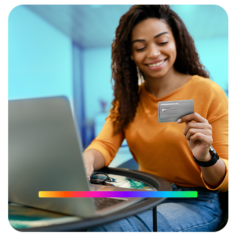 Jovem negra, sorrindo, de camiseta laranja e calça jeans, segurando um cartão do Magazine Luíza, enquanto com a outra mão, digita em um notebook em cima de uma pequena mesa. 