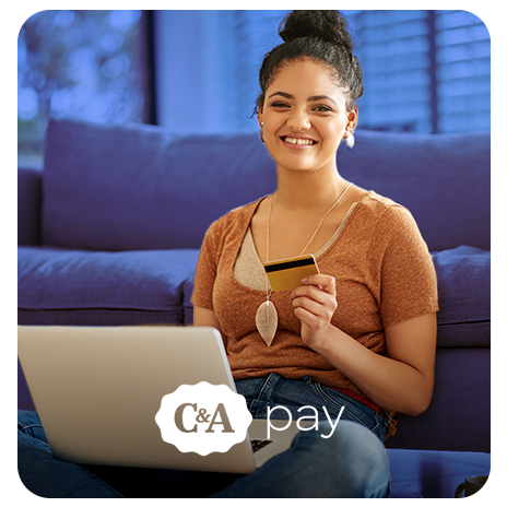 Mulher jovem, sorrindo, sentada no chão da sala de casa, com notebook no colo, encostada em seu sofá, segurando um cartão de crédito.