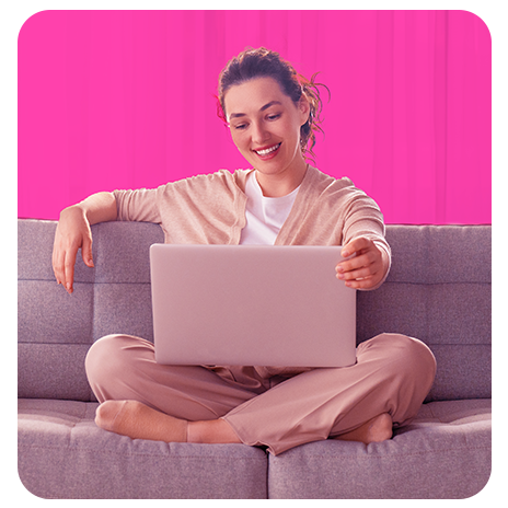 Mulher jovem, sorrindo, vestindo um conjunto bege, sentada de pernas cruzadas em um sofá cinza, com um notebook no colo, com um fundo rosa por trás.