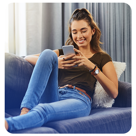 Mulher jovem, branca, sorrindo, de jeans e blusinha regata marrom, sentada em um sofá azul em sua casa, olhando o celular.