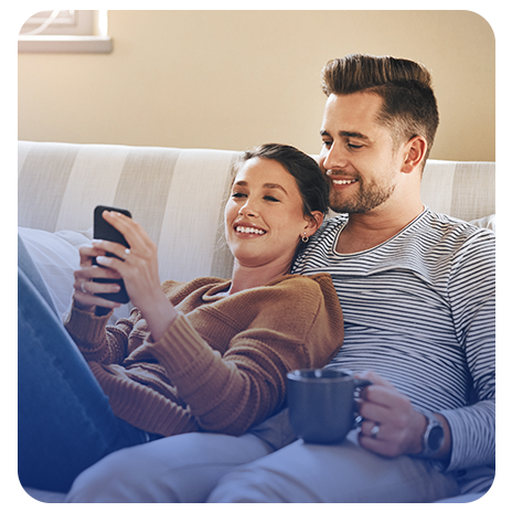 Um casal de jovens, brancos, sorrindo, sentados no sofá de casa, ele segurando uma caneca e ela segurando e olhando o celular.